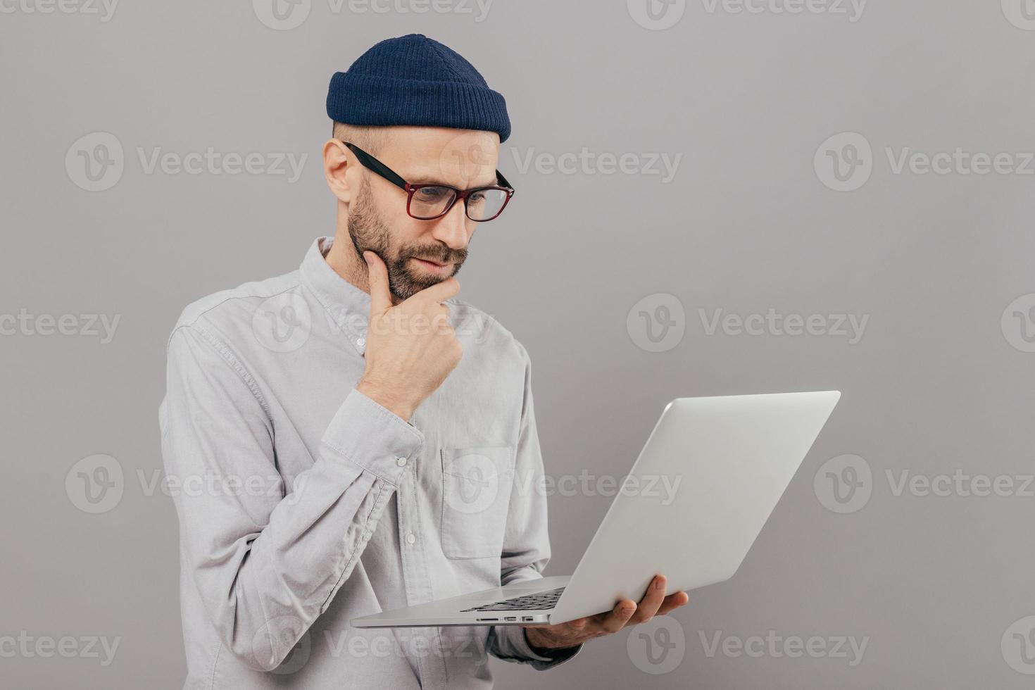 Attentive student holds chin, focused in monitor of laptop computer, searches information for project, connected to wireless internet, isolated over grey studio wall. Networking and people concept photo