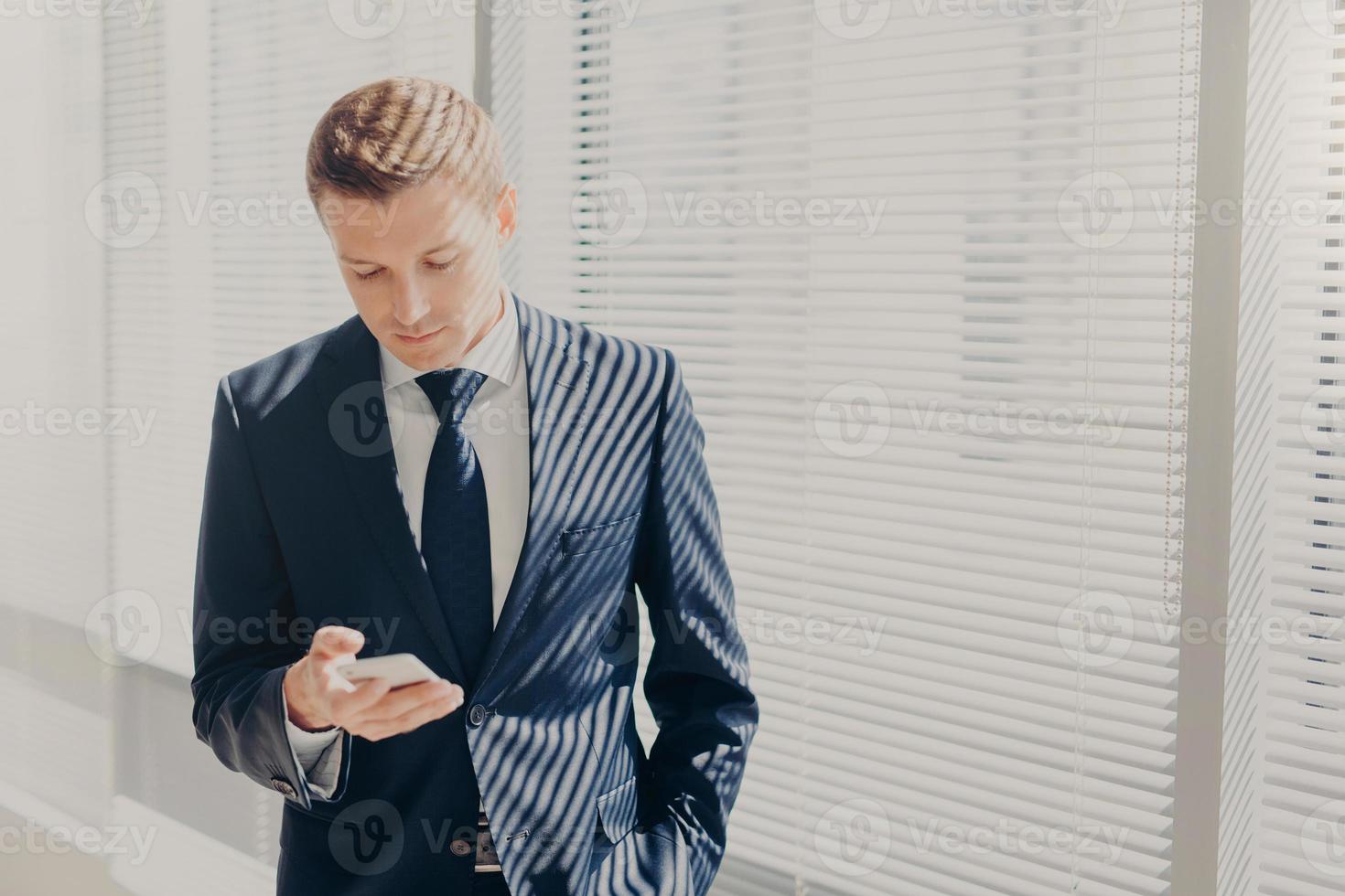 Good looking male executive dressed in corporate clothing, types text message, stands in office building, types feedback on mobile phone, stands near window at his cabinet. Technology concept photo