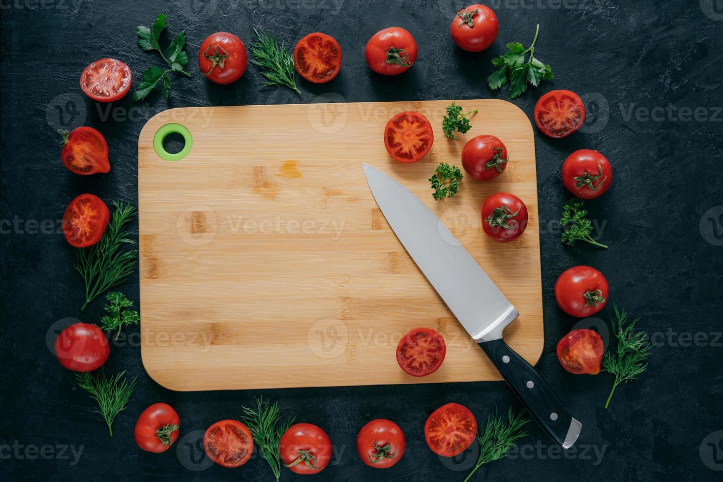 Tomatoes frame around wooden cutting board. Ripe vegetables and slice, green parsley and dill near kitchen board and knife. Food art photo