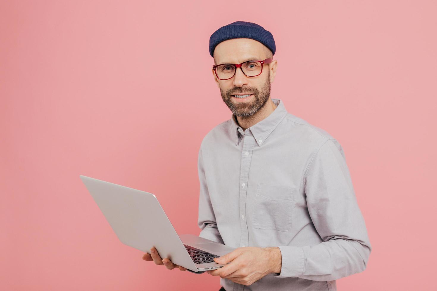 periodista masculino talentoso con una apariencia atractiva, tiene una computadora portátil moderna, trabaja en un nuevo artículo, vestido con ropa de moda, modelos de fondo rosa, conectado a internet inalámbrico foto