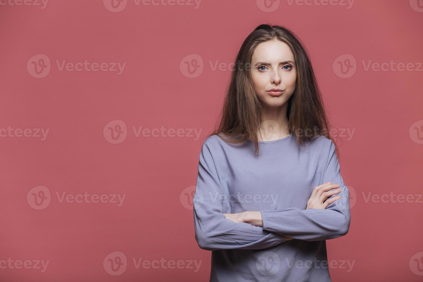 Confident serious attractive female model keeps hands folded, wears casual clothing, listens attentively information, poses against pink background with blank copy space for your promotional text photo