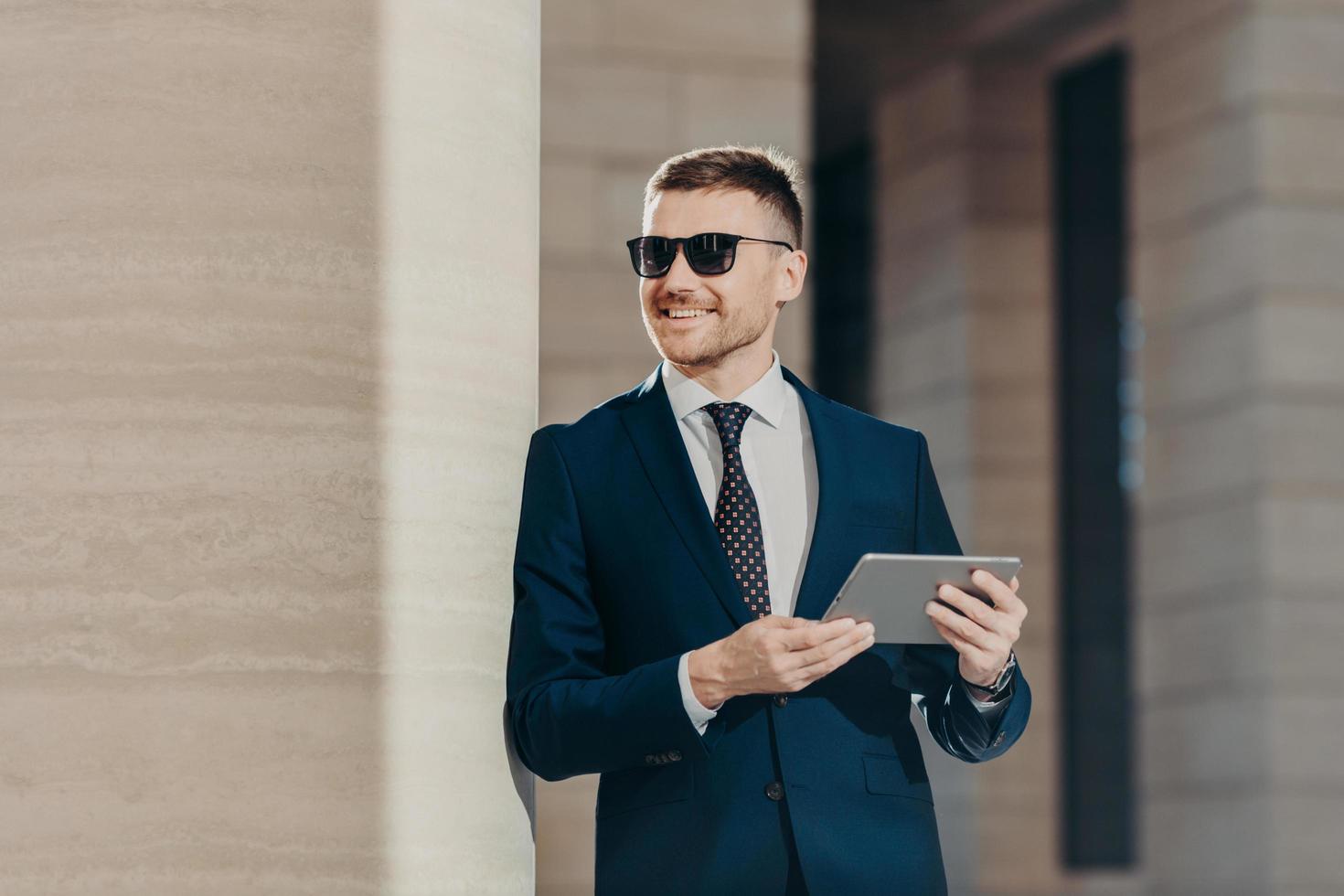 Portrait of handsome young male employer holds digital tablet, connected to wireless internet, wears sunglasses and formal suit, has free time after hard working day. People and career concept photo