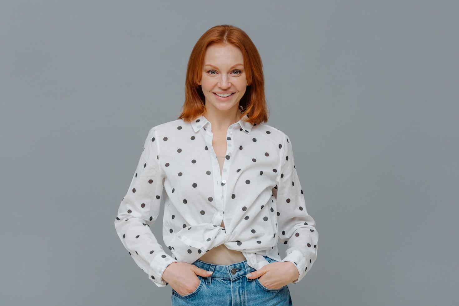 Fashionable positive woman wears polka dot shirt and jeans, keeps hands in pockets, stands self assured against grey background, has happy casual talk with interlocutor. Emotions, fashion concept photo