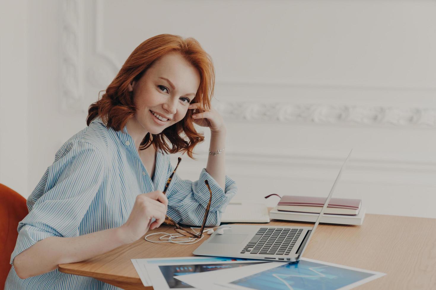 Satisfied businesswoman with foxy hair, checks accounting documentation in online database, works on modern laptop indoors, poses at workplace, surrounded with papers, thinks about creative ideas photo