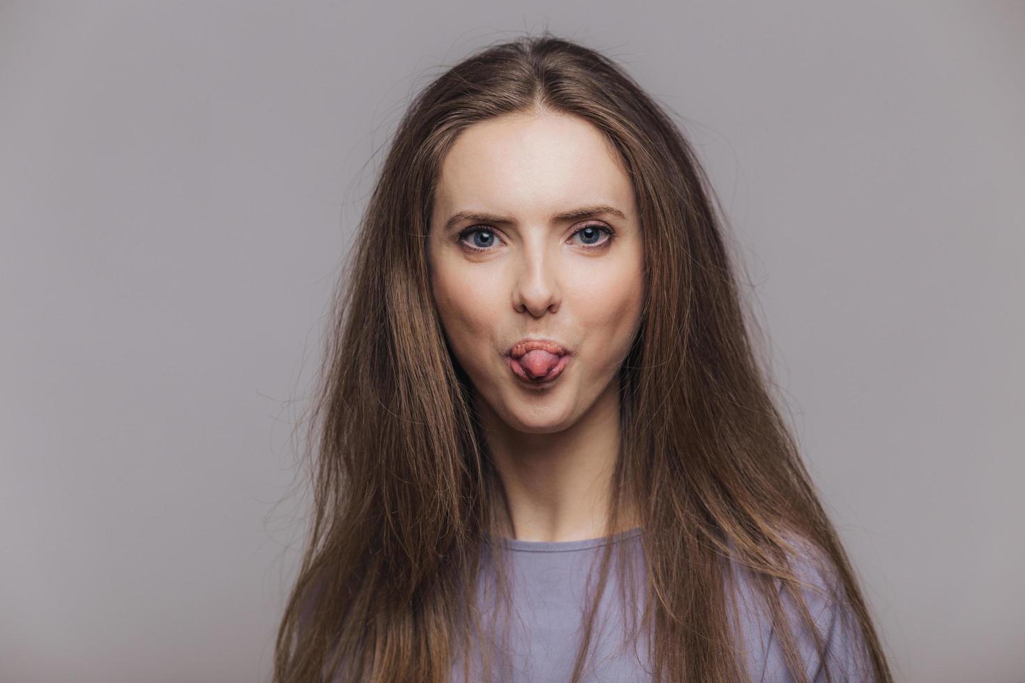 Funny brunette female with atractive look, shows tongue, makes grimace and has fun indoor, looks with blue eyes at camera, has dark straight hair, isolated over grey background. Positivity concept photo