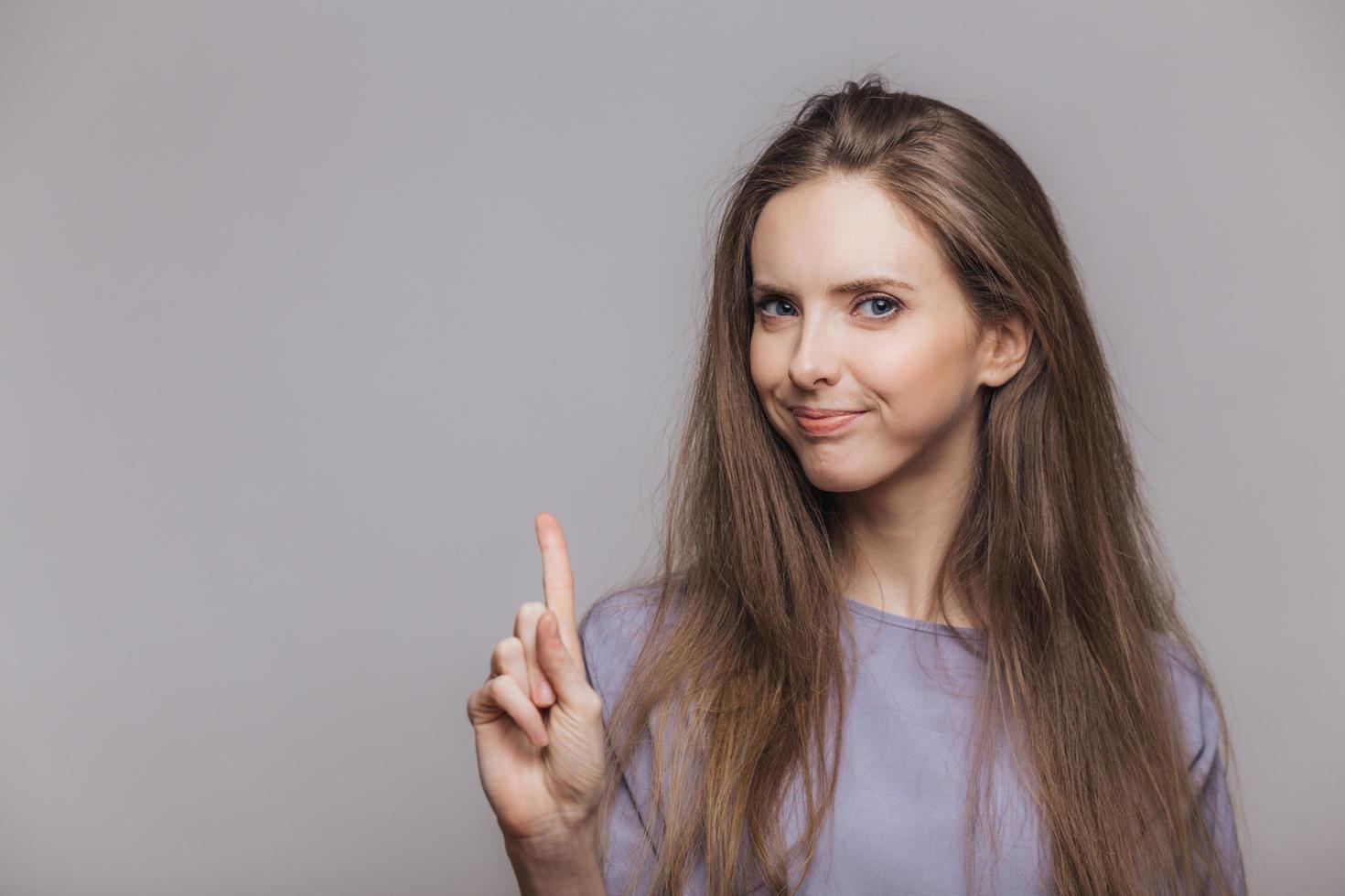 Horizontal portrait of pleasant looking brunette female model with dark hair, keeps fore finger raised upwards, shows something, poses against grey background with blank copy space for advertisment photo