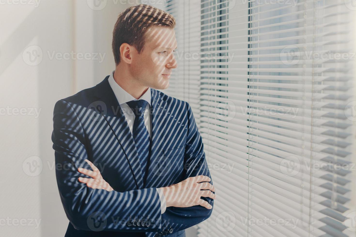 Contemplative male director keeps arms folded, thinks about future work plans, dreams about high profit of his company, looks thoughtfully through office window, dressed in formal elegant suit photo
