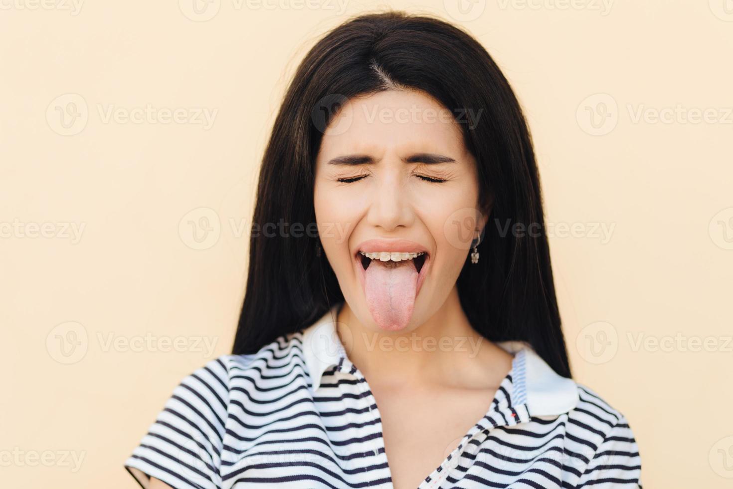 Portrait of beautiful woman makes grimace, keeps mouth wide opened and shows tongue, keeps eyes closed, wears casual clothes, poses against studio background. People and facial expressions concept. photo