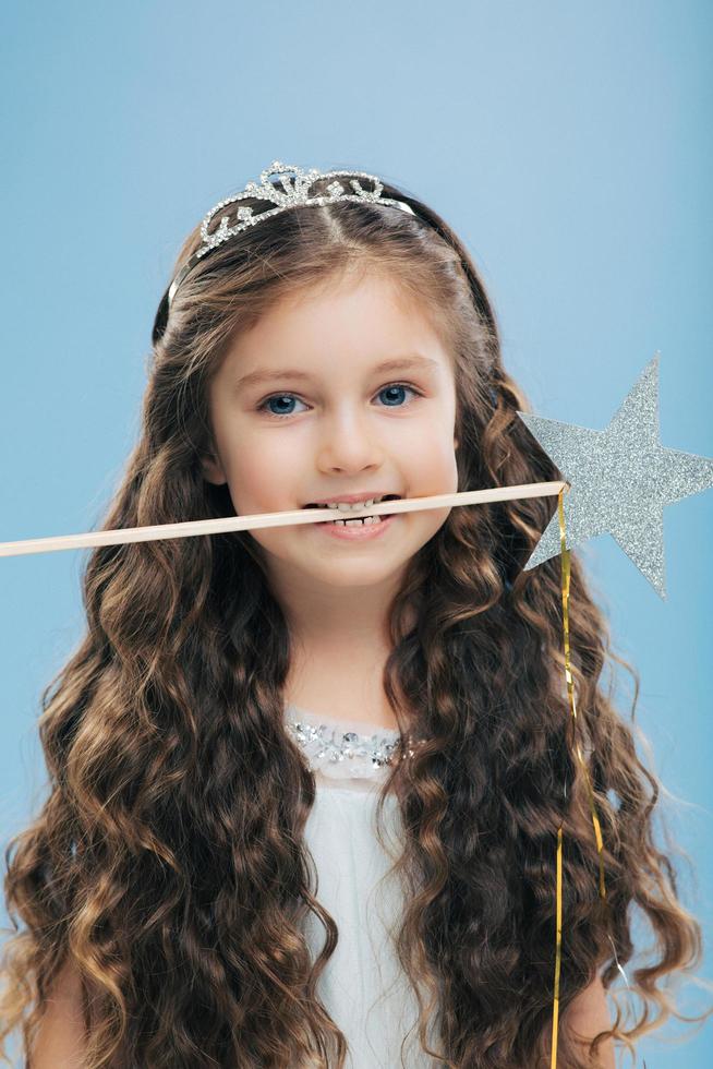 Photo of attractive woman kid with pleasant appearance, has long dark hair, holds magic wand in mouth, poses over blue background, dressed in princess dress. Vertical image. Childhood concept