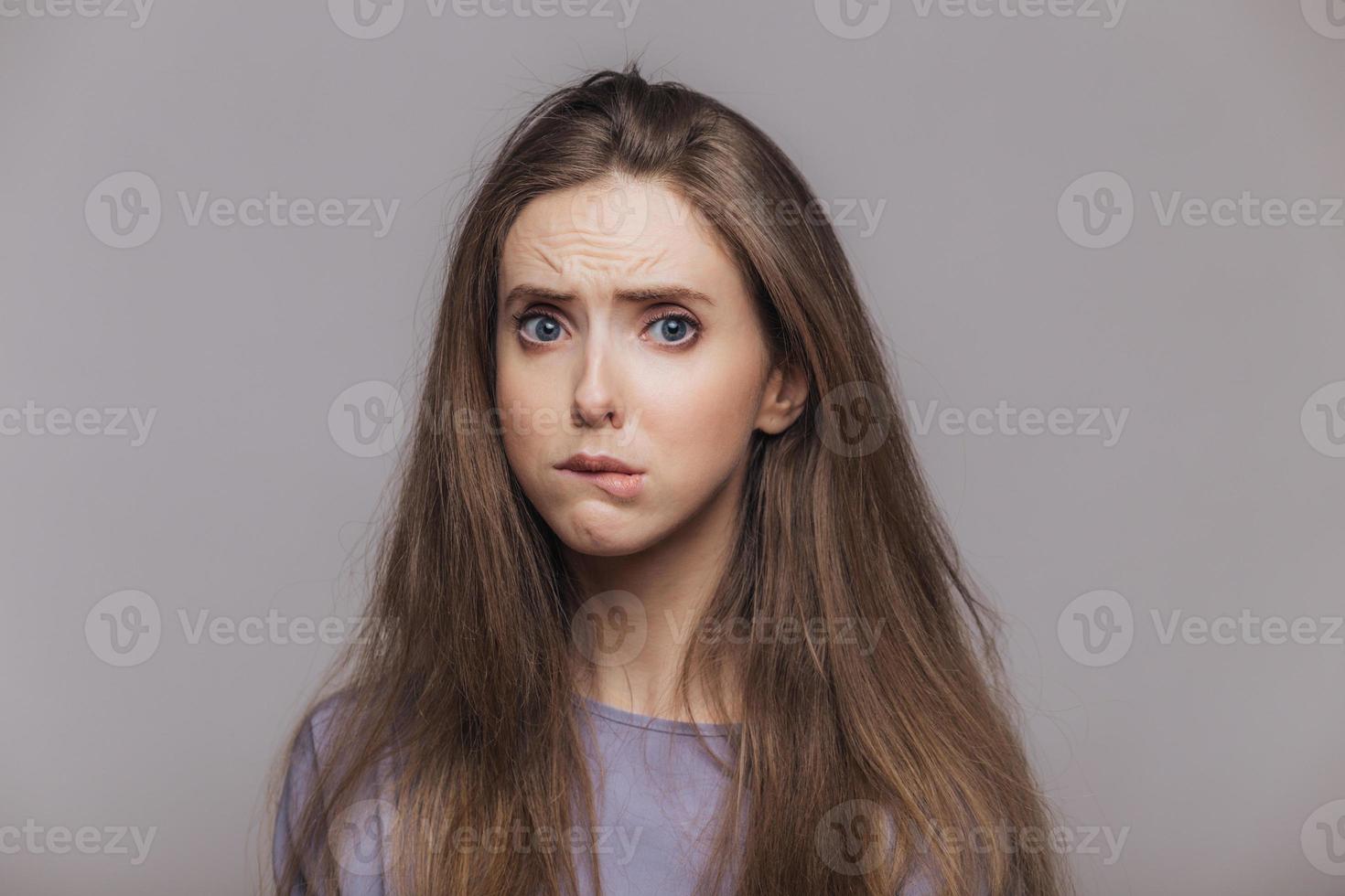 Offended stressful female bites lower lip with dissatisfaction, has sorrorful expression, expresses negative emotion, being in low spirit after quarrel with somebody, isolated over grey background photo