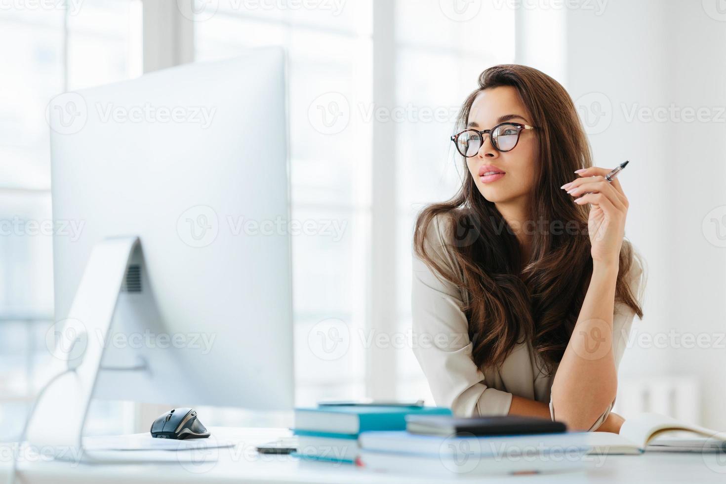 Dark haired lady focused into big monitor, sits at desktop, holds pen and writes notes, wears spectacles for vision correction, poses in coworking space. Femlae manager at workplace, makes research photo