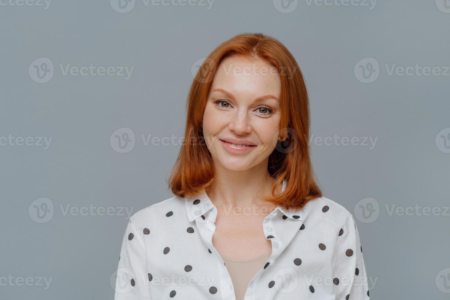 Portrait of attractive ginger European woman smiles gently at camera, wears makeup, dressed in polka dot blouse, looks directly at camera, models against grey background, thinks about changing job photo