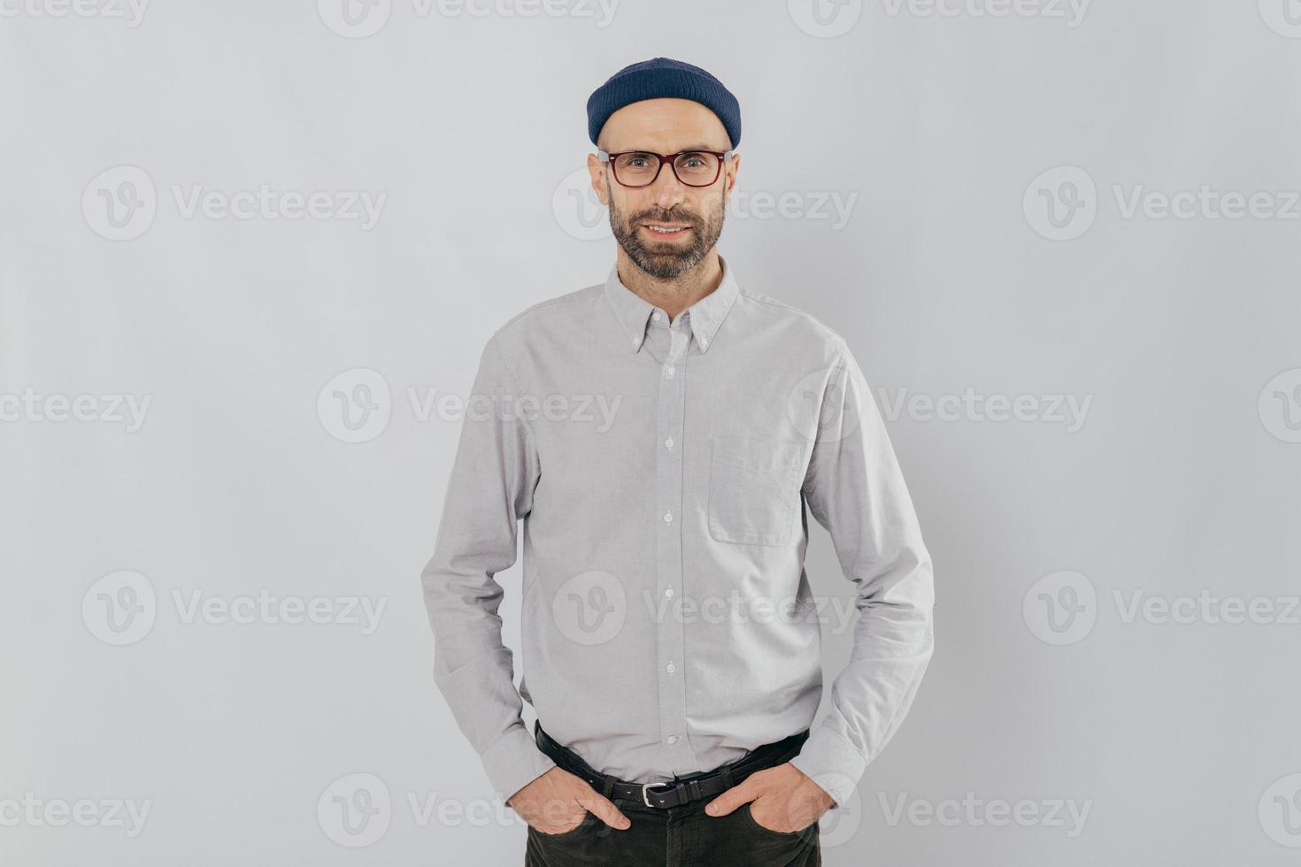 Indoor shot of satisfied Caucasian man in spectacles with bristle, keeps hands in pockets, dressed in formal stylish clothes, has gentle smile, happy to be promoted, isolated over white background. photo