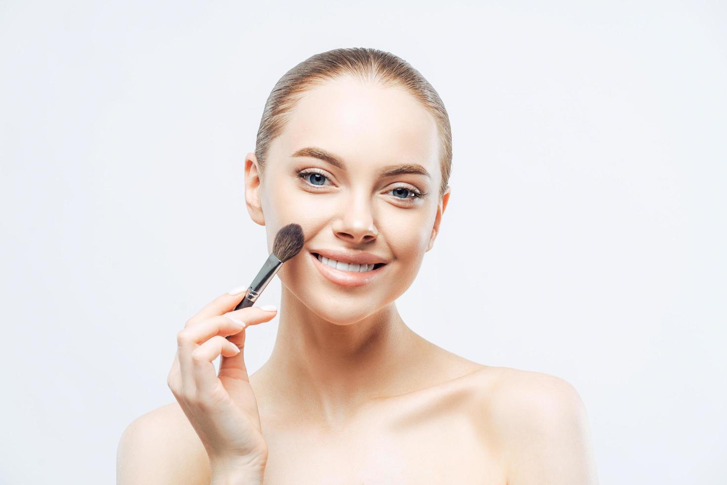 Adorable young brunette woman applies cosmetic tonal foundation with beauty brush, smiles tenderly, has healthy skin, well cared body, isolated on white background. Women, skin care, makeup concept photo