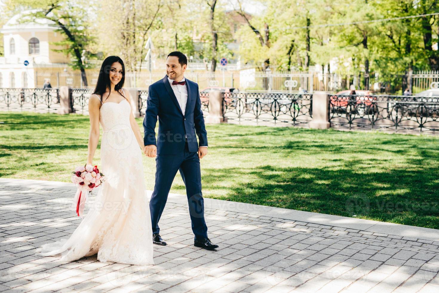 Outdoor portrit of lovely married couple walk together, keep hands together, enjoy nice weather. Smiling bride and bridegroom walk on path, have good mood. Lovely pair has romantic relationship photo