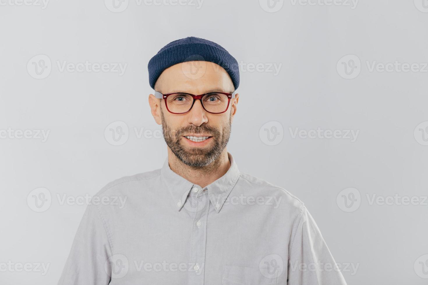 Horizontal view of pleased smiling man wears glasses, hat and shirt, being satisfied with good news, models against white background. Unshaven hipster rejoices new purchase. Fashion concept. photo