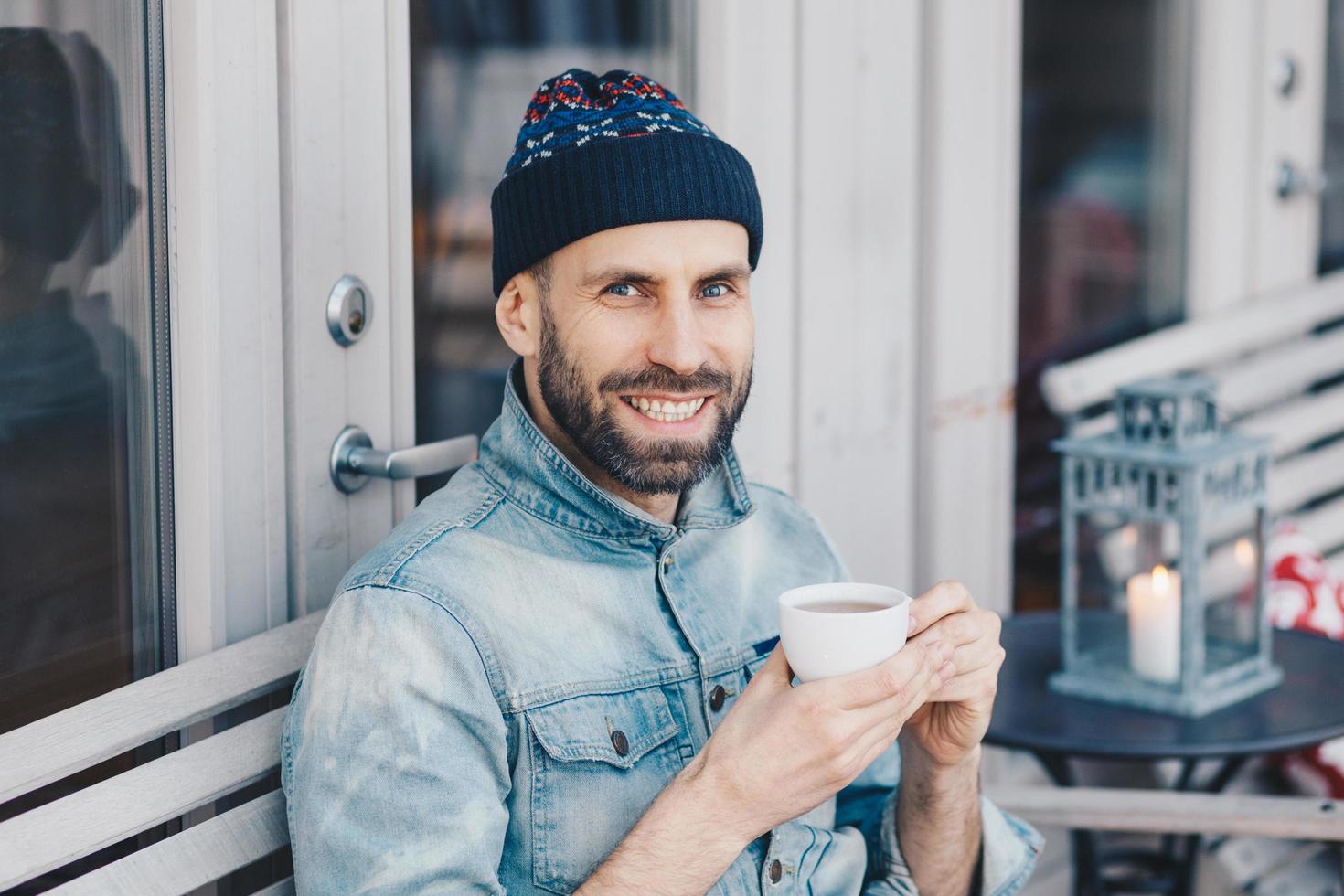 Cheerful bearded male in denim jacket, has positive happy expression, spends free time at terrace, drinks aromatic tea, feels relaxed and delighted. People, positive emotions and recreation time photo