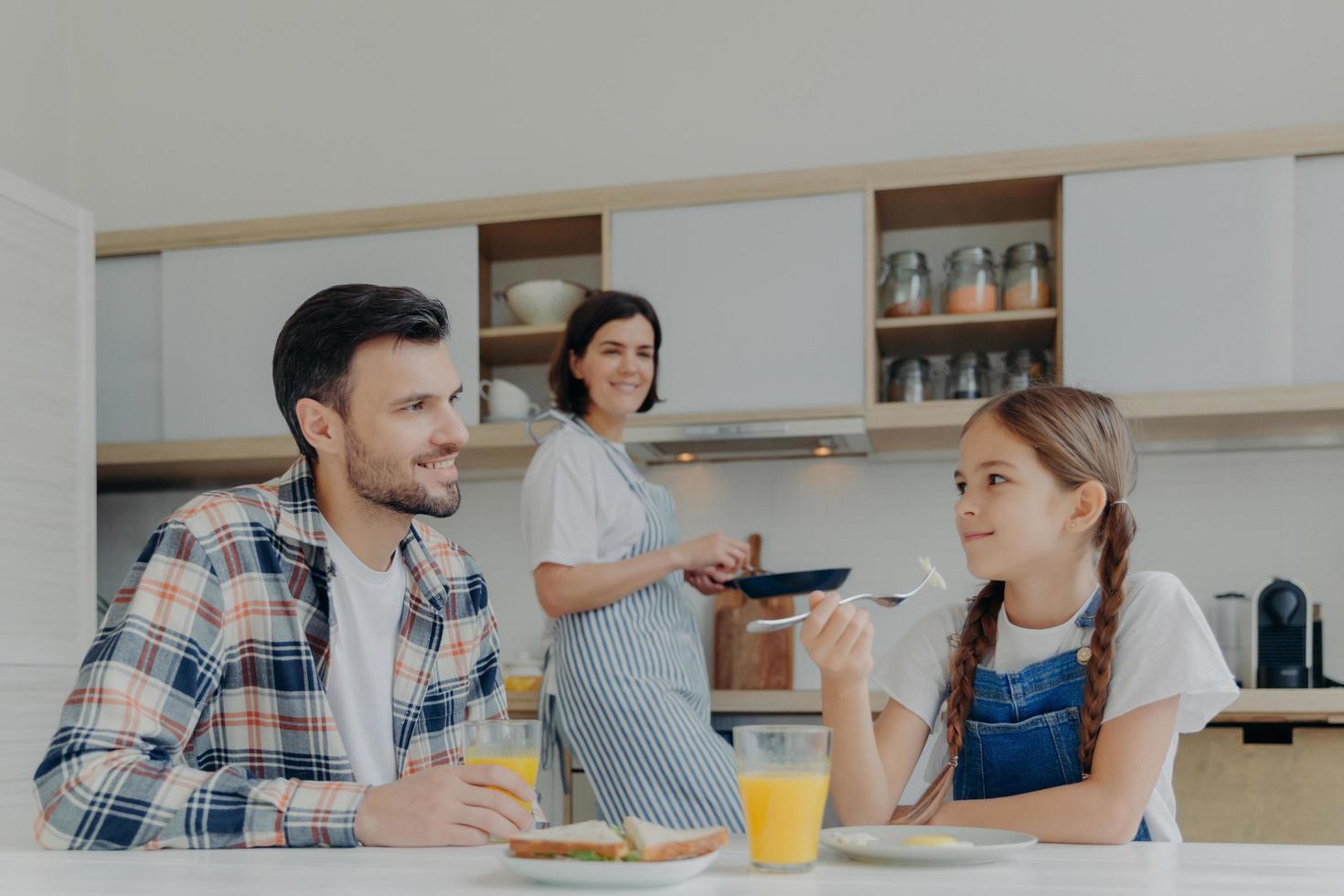 Happy family have breakfast together in kitchen. Positive little child talks with father while eat delicous dish, mother stands in background near stove, holds frying pan, busy with cooking. photo