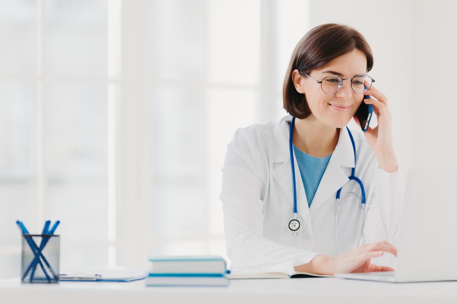 Smiling therapist focused at screen of laptop computer, calls via modern smartphone, wears medical gown, transparent glasses, sits at coworking space, surfes internet. Copy space for your information photo