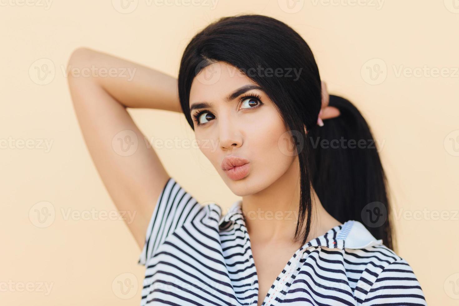 Cute young woman has dreamy expression, keeps lips round, has make up, makes hair in pony tail, dreams about something, looks thoughtfully, isolated over beige background. Youth and beauty concept photo