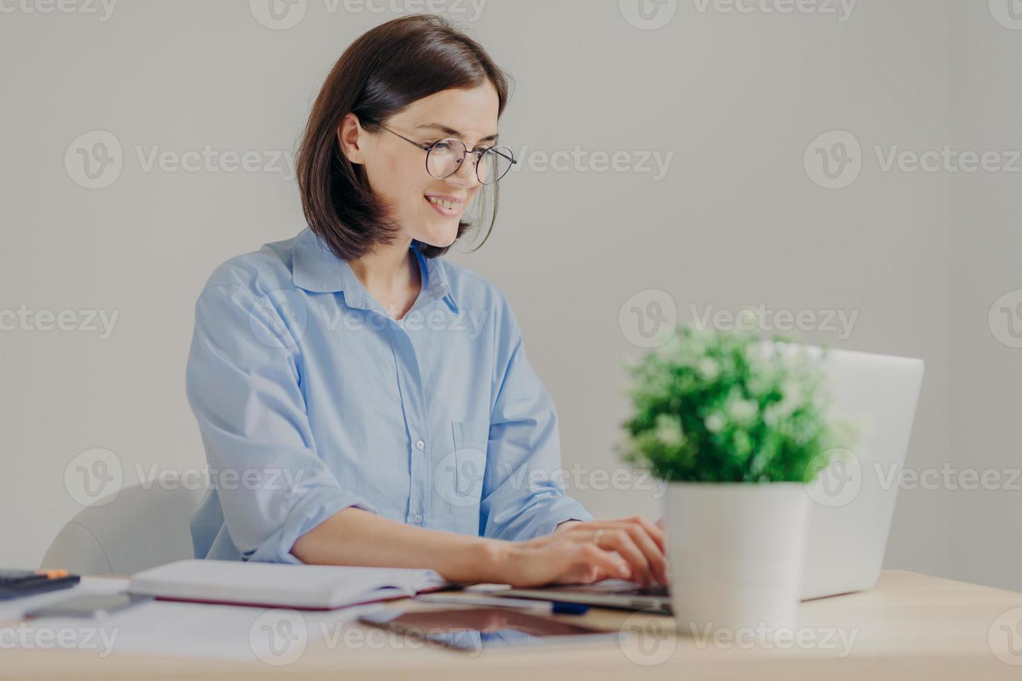 Cheerful successful female freelnacer works remotely, makes research, keyboards on laptop computer, surfes in internet, uses free internet connection. People, business and occupation concept photo