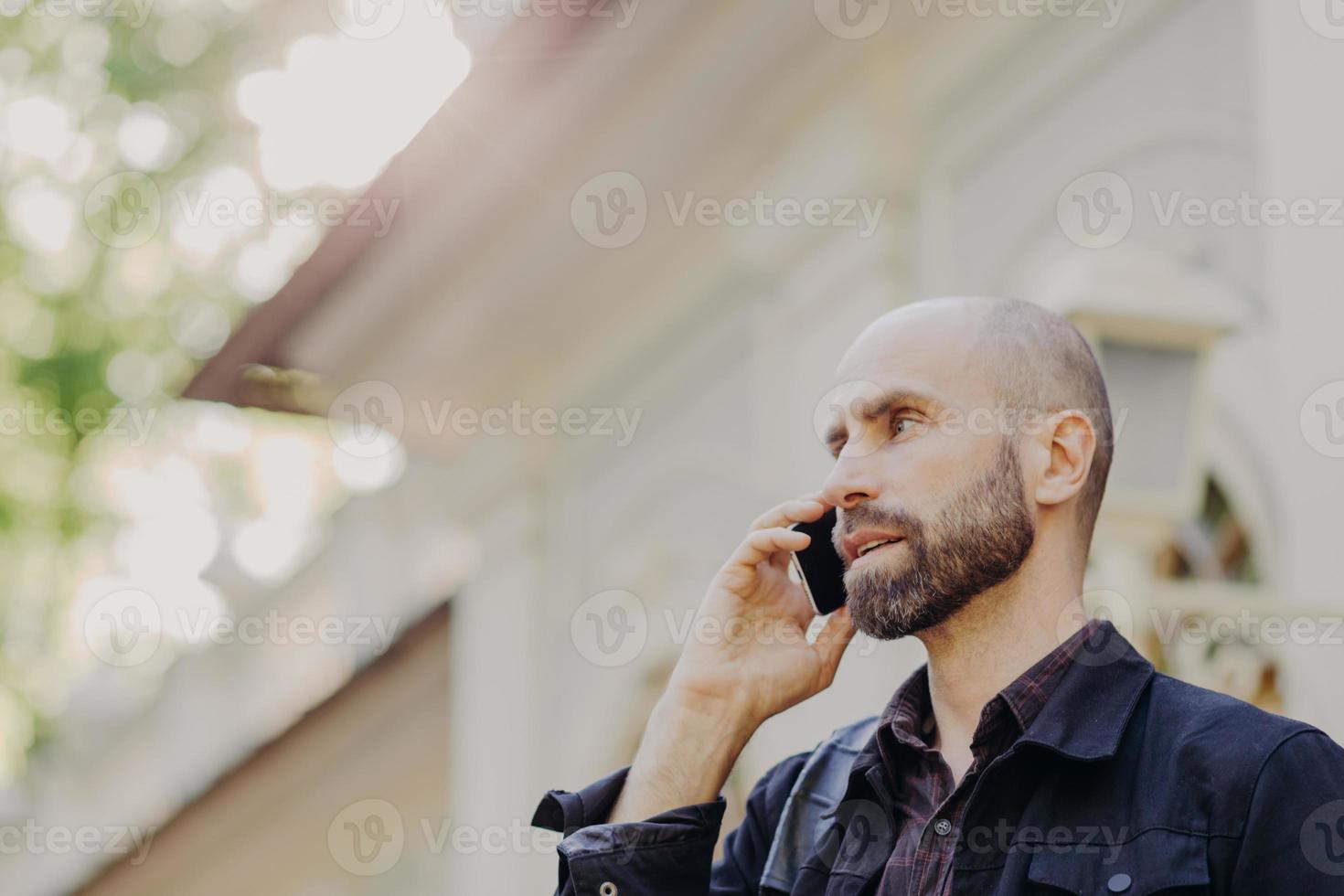 vista desde arriba de un atractivo hombre de mediana edad con barba, habla con un socio comercial a través de un teléfono inteligente, posa contra un fondo borroso, disfruta usando tecnologías modernas. el hombre tiene conversacion foto