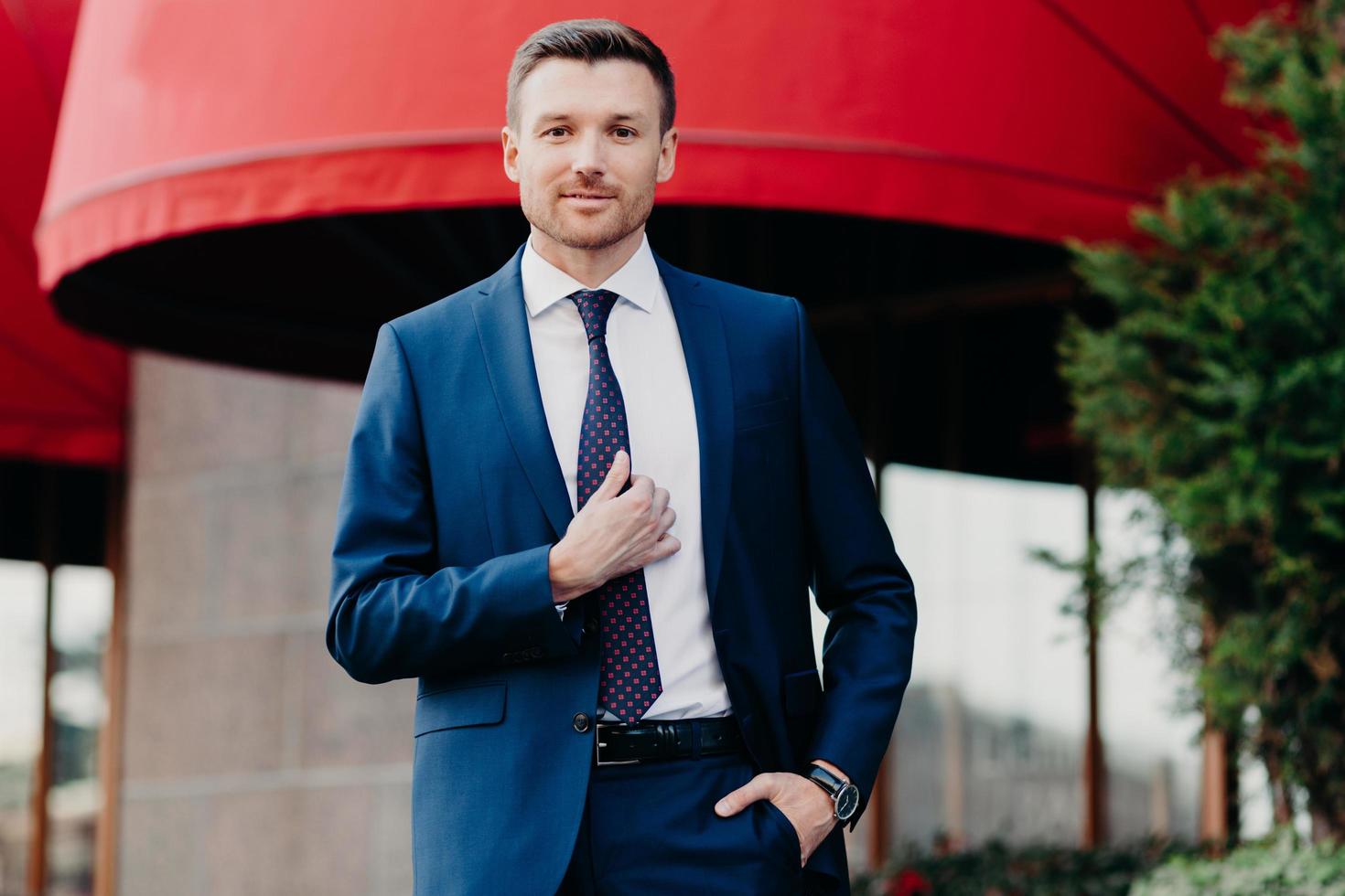 Handsome male company owner dressed formally, stands outside, has confident expression, ready for business meeting. Successful young attractive executive manager poses outdoor near office building photo