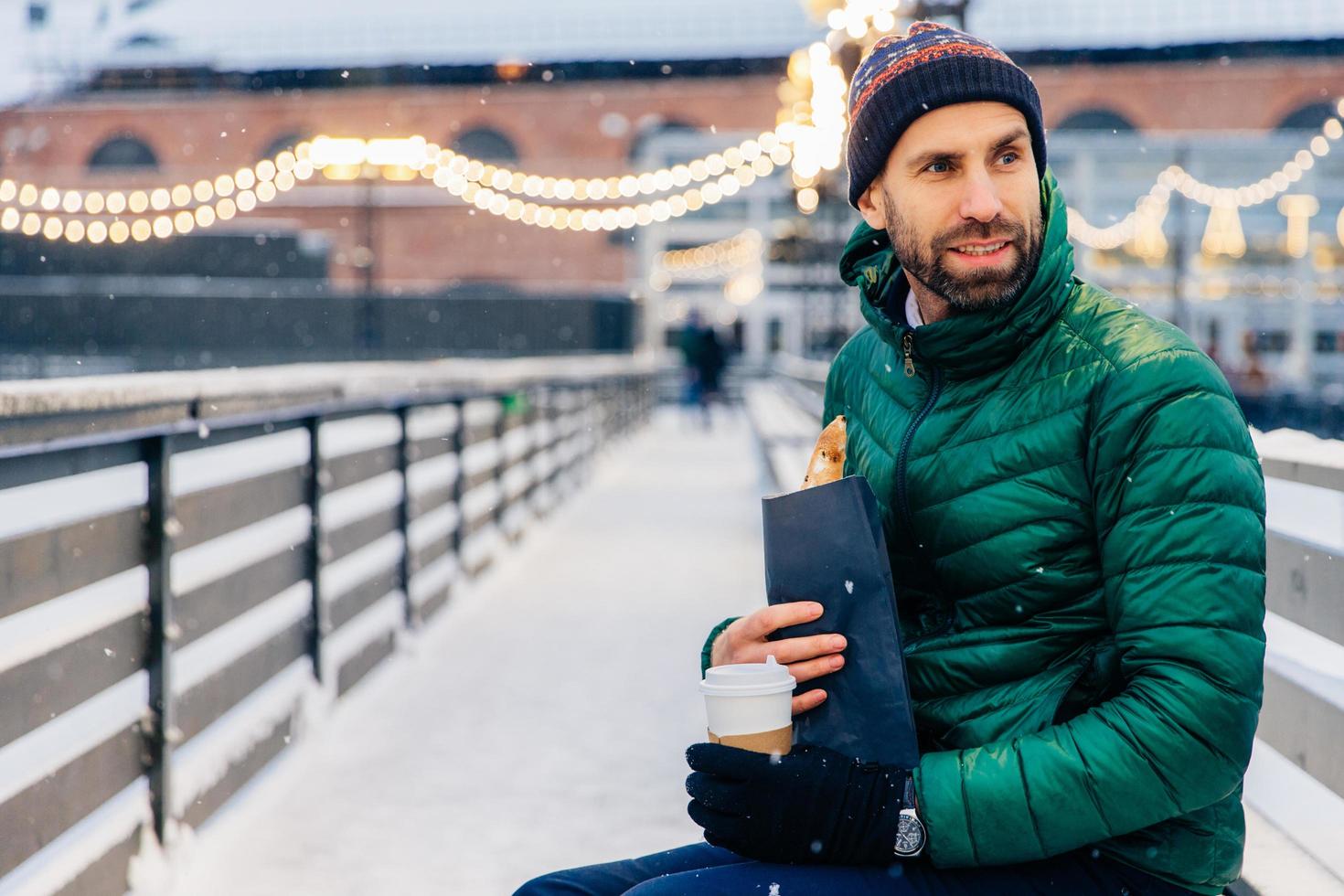 Middle aged man looks thoughtfully aside, has snack outdoors during winter weather, drinks hot coffee and eats roll, walks on street, enjoys snow. People, lifestyle, rest, lunch concept photo