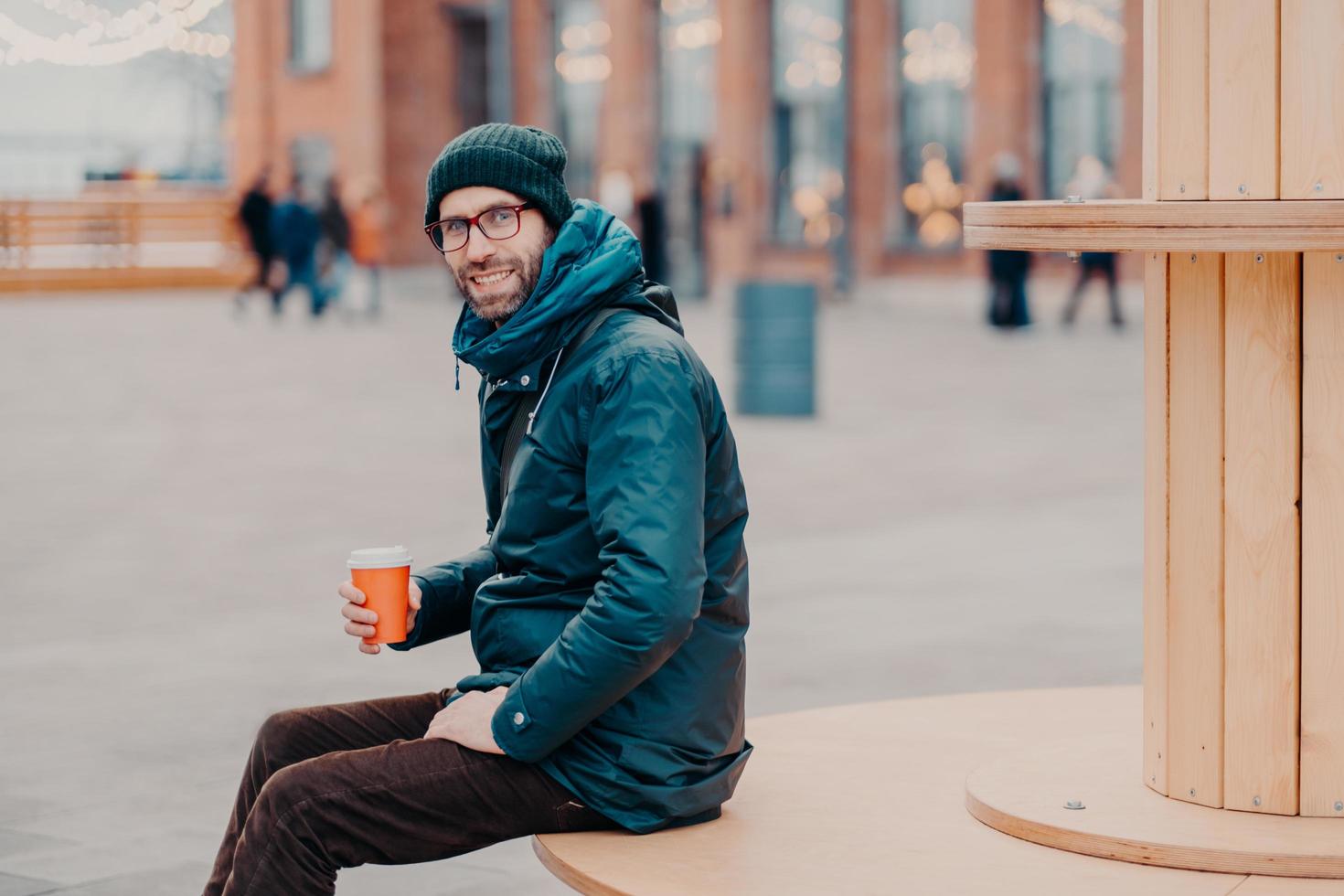 vista horizontal de un alegre hombre europeo con cerdas gruesas, usa sombrero y chaqueta, sostiene una taza de café desechable, respira aire fresco, posa en la calle. modelo masculino disfruta de una bebida fresca afuera foto