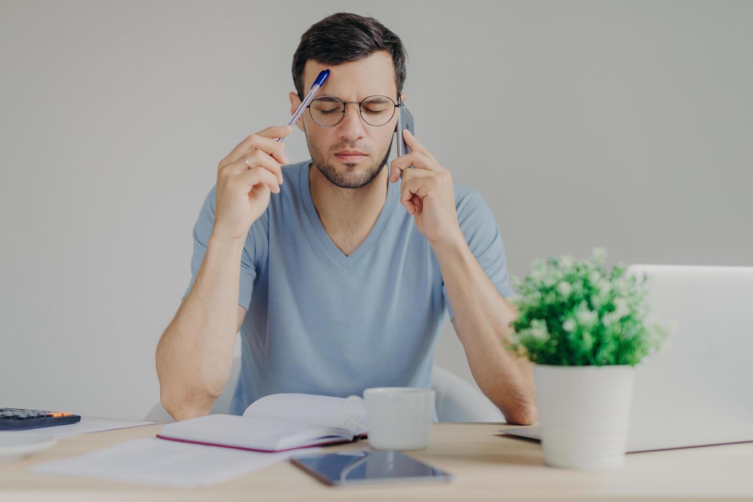 Concentrated young male manager makes internet project, has phone conversation, browses information on laptop computer, tries to thinks what write, isolated over grey background. Job concept photo