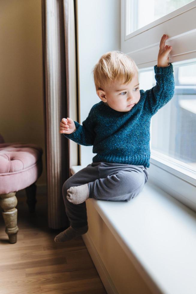 Naughty small kid with blonde hair and blue eyes, sits on window sill, looks out of window, notices something in yard. Curious cute child plays alone, looks outside with attentive expression photo