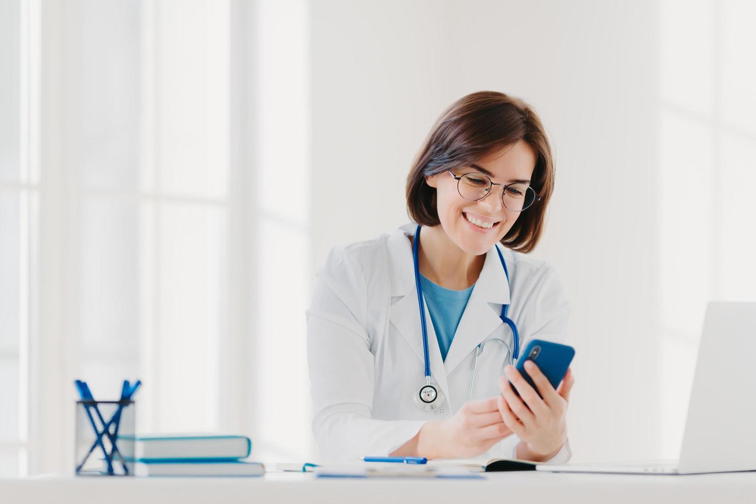 Positive female medical worker involved in chatting, holds mobile phone and sends messages, wears medical gown, has friendly cheerful expression, poses at desktop gives online consultation for patient photo