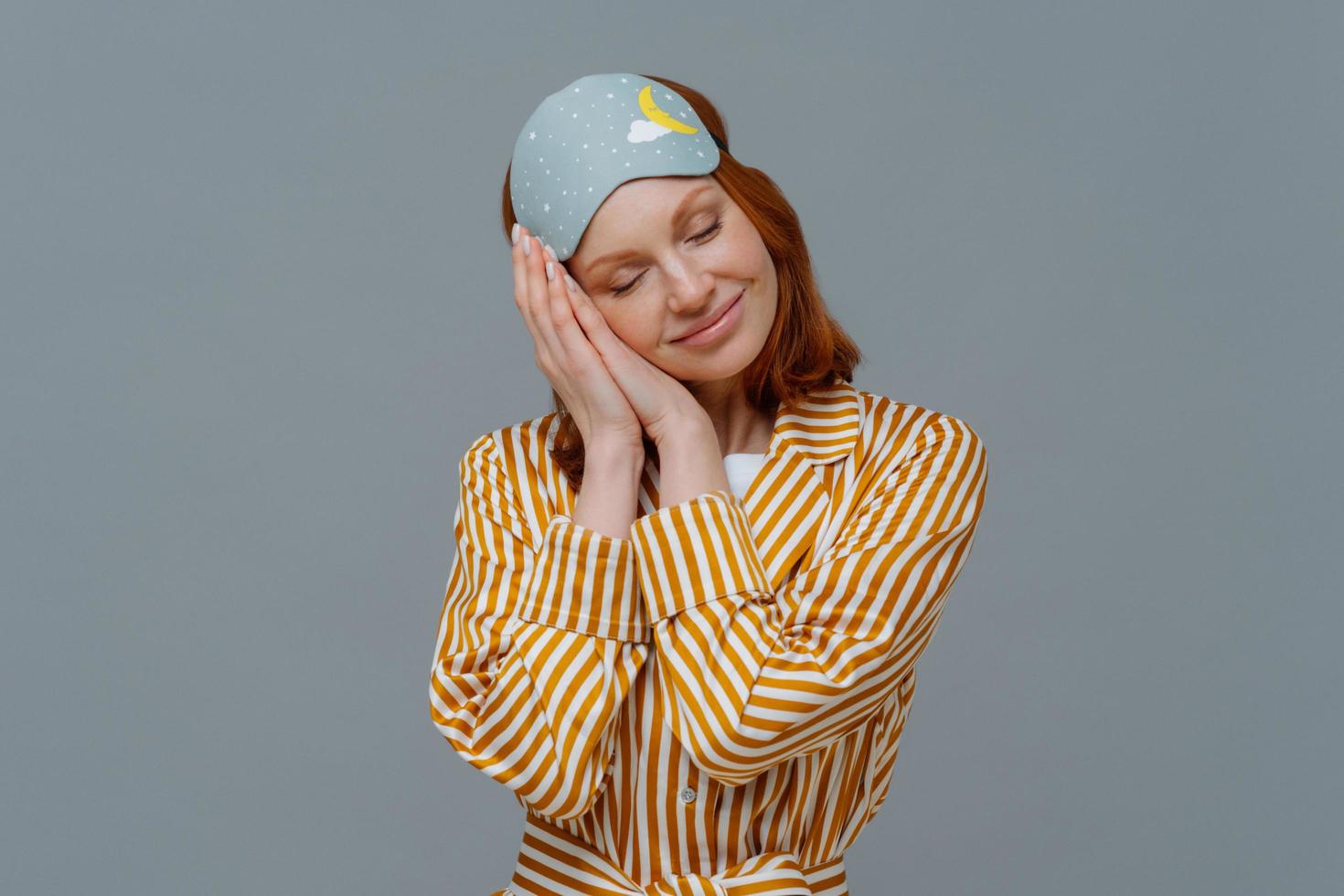 Pleased cheerful woman wears blindfold and striped pajamas, has happy expression, awakes in good mood, has healthy sleep habits, feels totally relaxed, smiles broadly, isolated on grey background photo