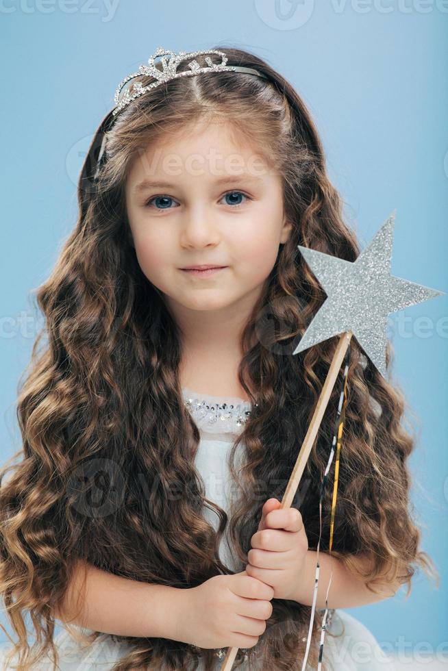Small kid angel has long crisp hair, blue eyes, holds magic wand in form of star, poses over blue background, has appealing appearance. Little princess waits for miracle, believes in fairy tale photo