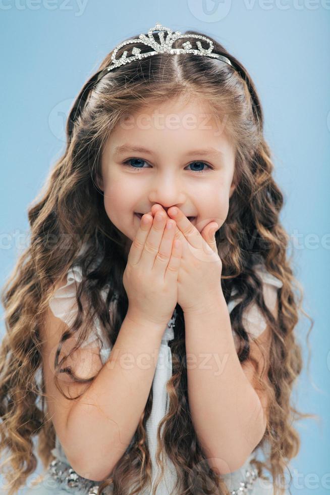 Vertical shot of positive small child covers mouth with hands, giggles positively, wears crown, expresses happiness, isolated over blue background, hears something funny. Children and emotions photo