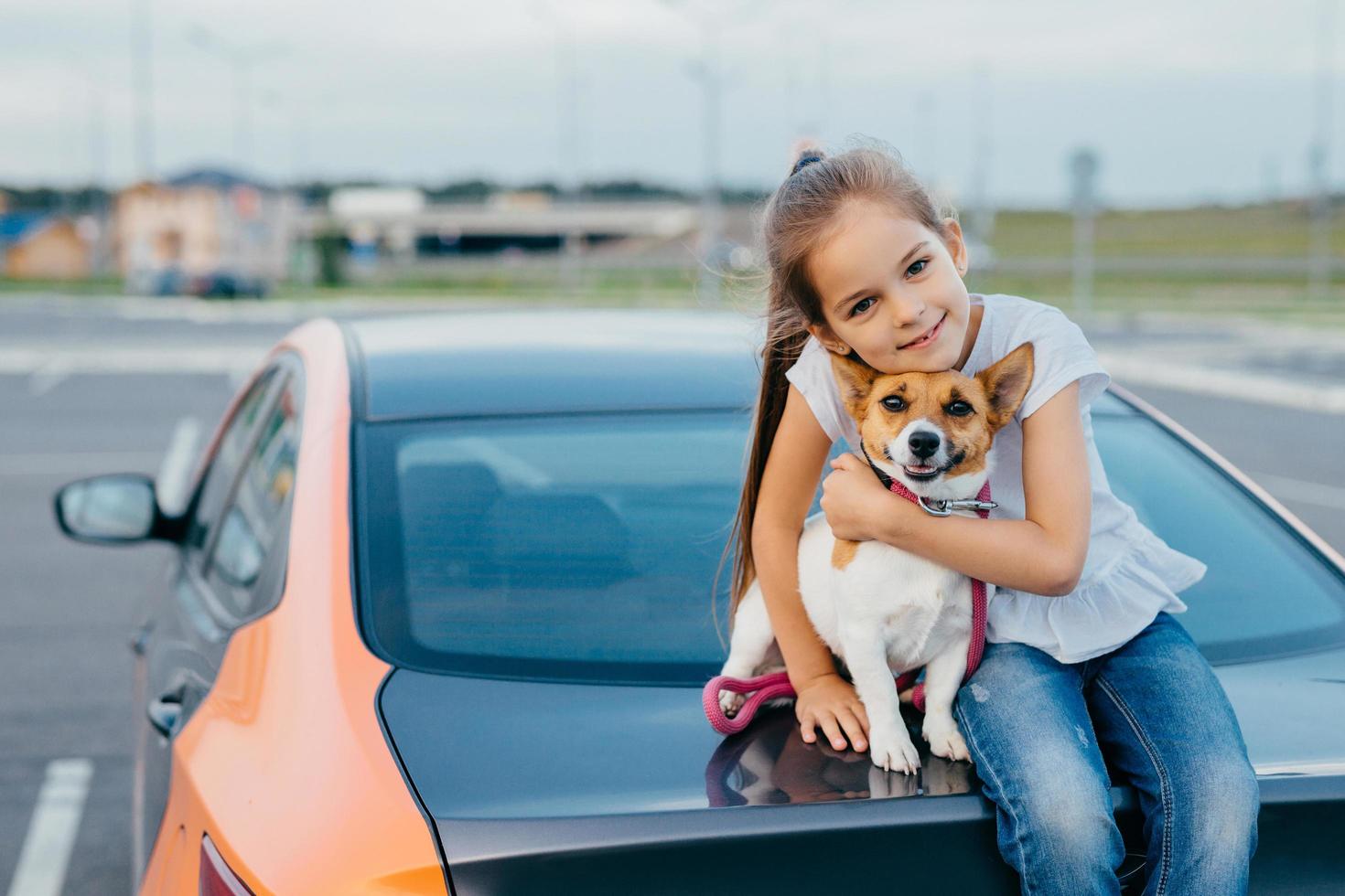 Small attractive female child embraces her favourite dog, sit together at trunk of car, have rest after stroll, enjoy summer day, have friendly relationship. Children, pets and lifestyle concept. photo