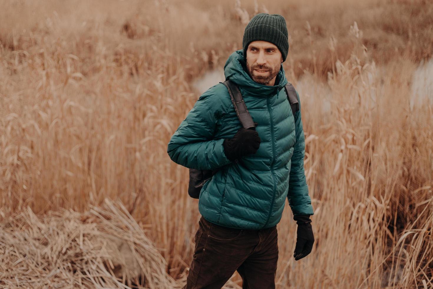 personas y concepto de viaje. foto de un hombre guapo vestido con una chaqueta abrigada y un casco, camina afuera, mira atentamente a un lado, posa contra el fondo del campo con espacio para copiar en el lado izquierdo para el texto