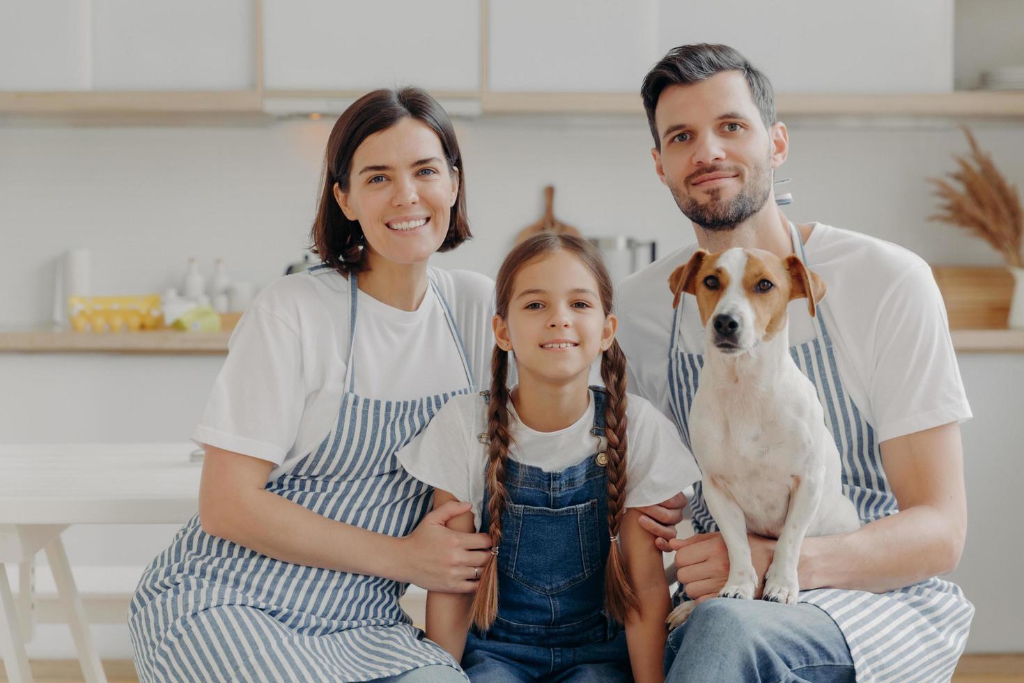 Family portrait of father, mother, daughter and pedigree dog pose together for making memorable photo, have rest after preparing dinner, poses against kitchen interior, have happy expressions photo
