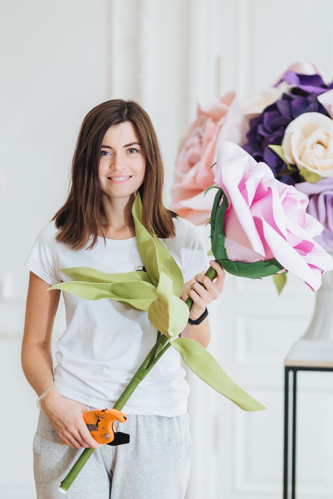 Vertical shot of professional young female designer or florist holds big bouquet, uses garden pinceps, dressed in casual outfit, likes decoration, owns business flower shop, makes arrangements. photo