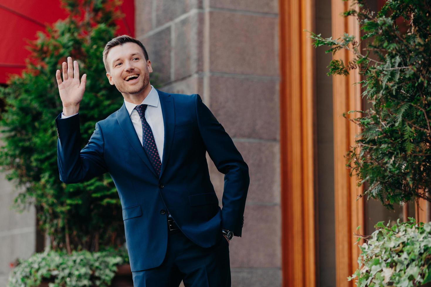 Shot of cheerful male banker in luxury black suit, waves with hand as notices friend on street, stands near his office, comes on work in good mood, has positive expression. People, occupation concept photo