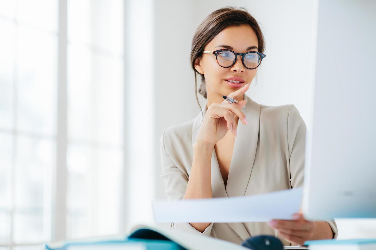 Female economist poses in coworking space, focused in monitor, verifies or checks figures from financial report, holds paper document and pen, has dark combed hair, wears formal business suit photo