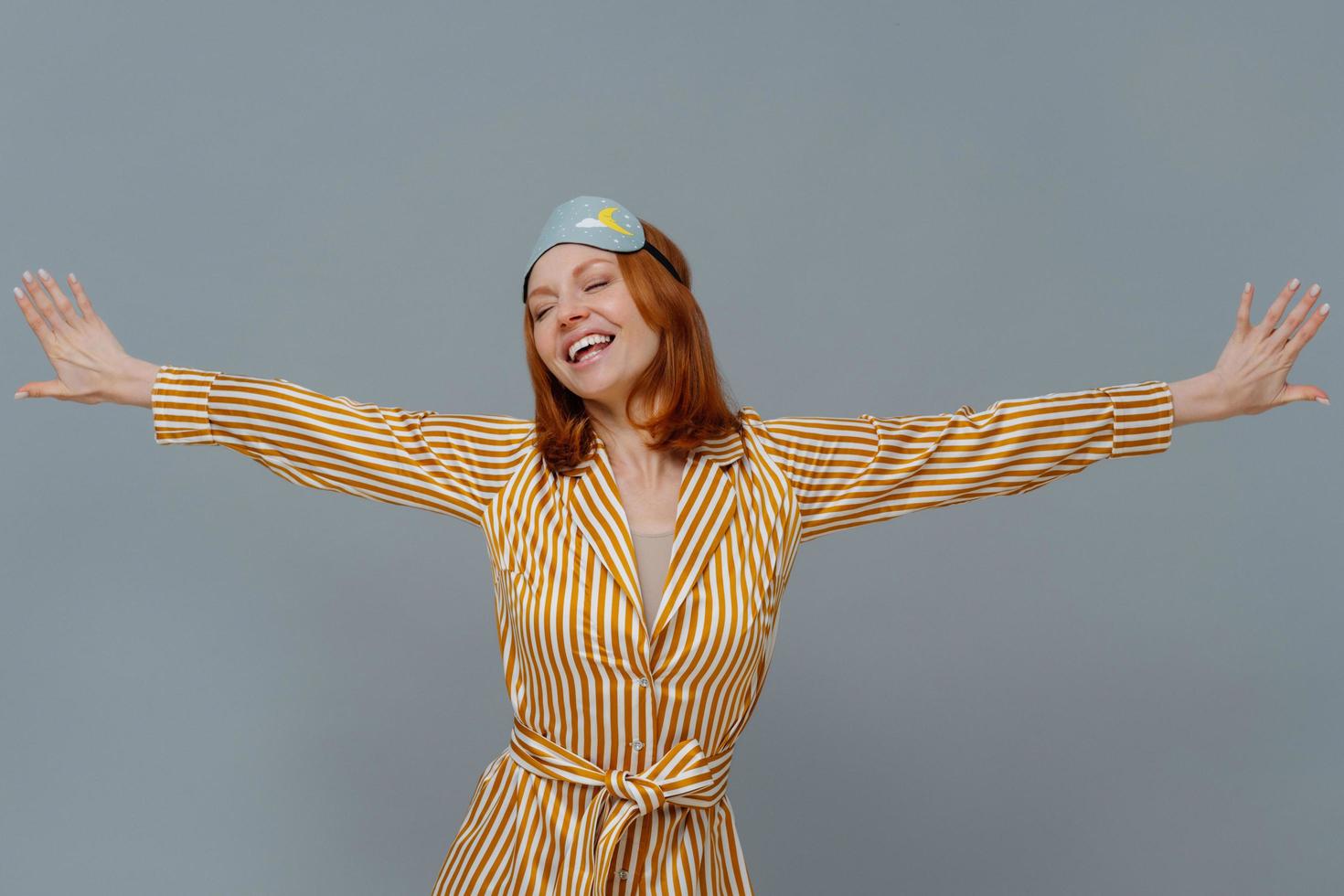 Positive relaxed woman wears blindfold and striped robe stretches arms sideways, smiles broadly, closes eyes, isolated over grey background, wakes up in bedroom, slept well. Good morning concept photo