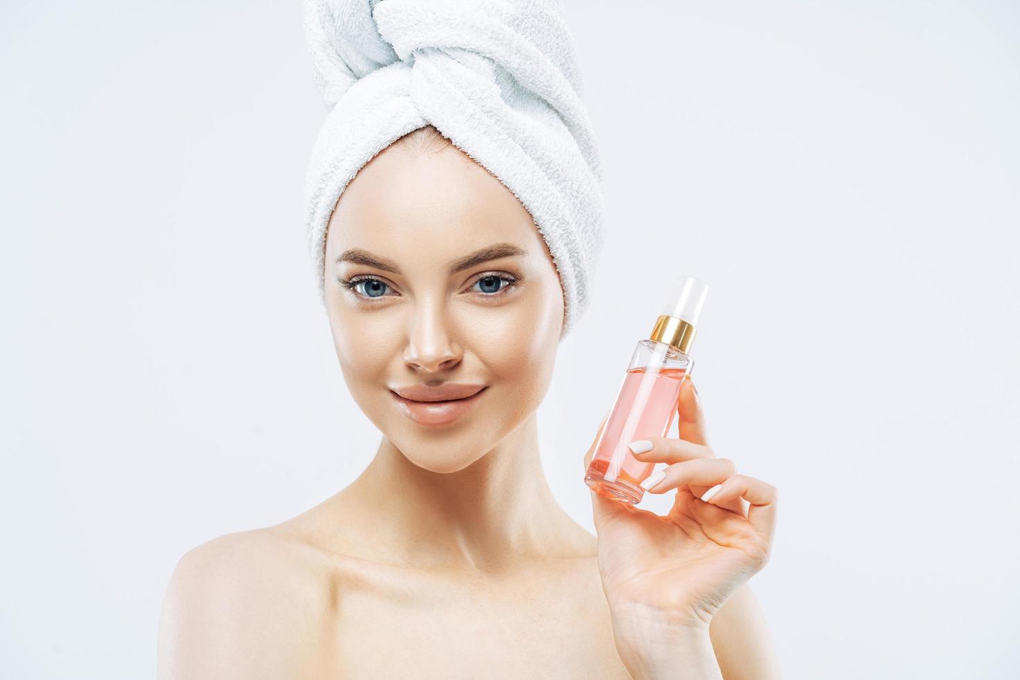 Young pleasant looking woman uses perfum, likes new smell, stands delighted indoor, applies makeup, has healthy skin wears bath towel isolated on white background has glamorous look. Pleasant smelling photo