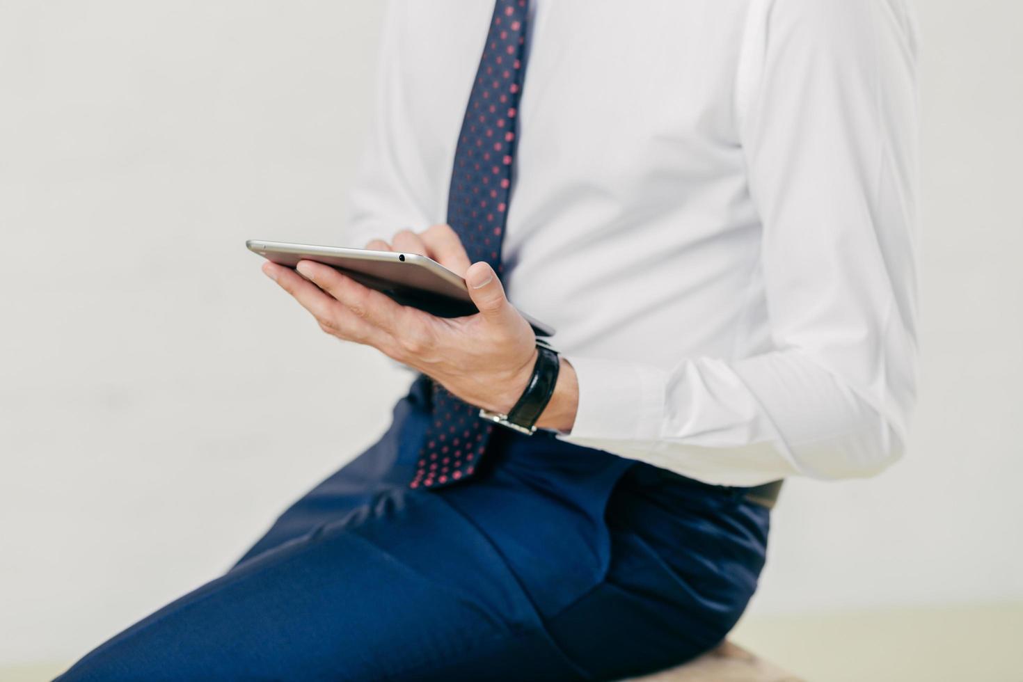 Cropped image of successful prosperous businessman in white shirt, black trousers, tie, holds modern tablet, checks notification, connected to wireless internet, isolated over white background photo