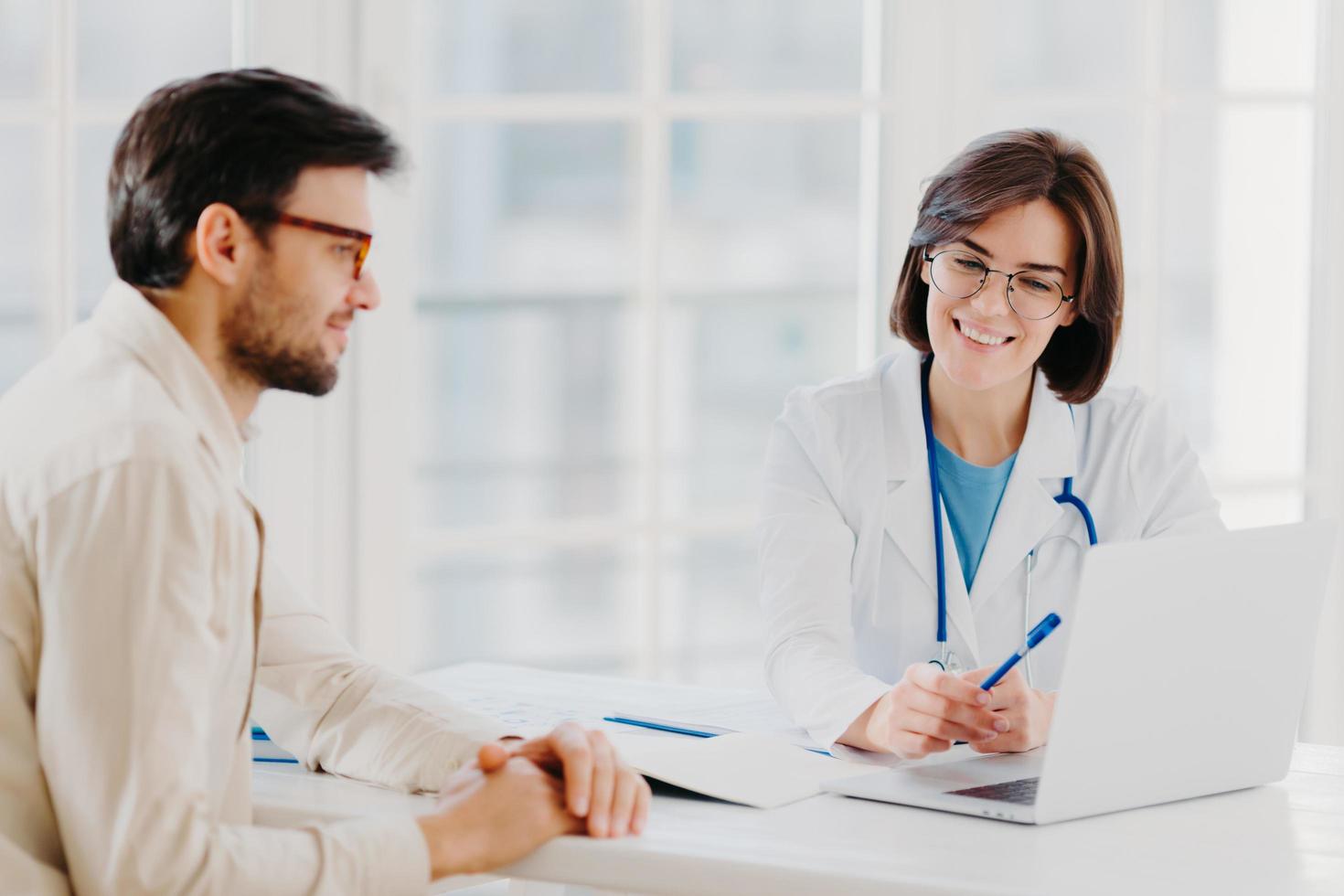 Caring woman doctor gives prescriptions to patient, sit together at table and look at laptop computer, fill up application form online. Sick man visit healthcare clinic, gets medical consultancy photo