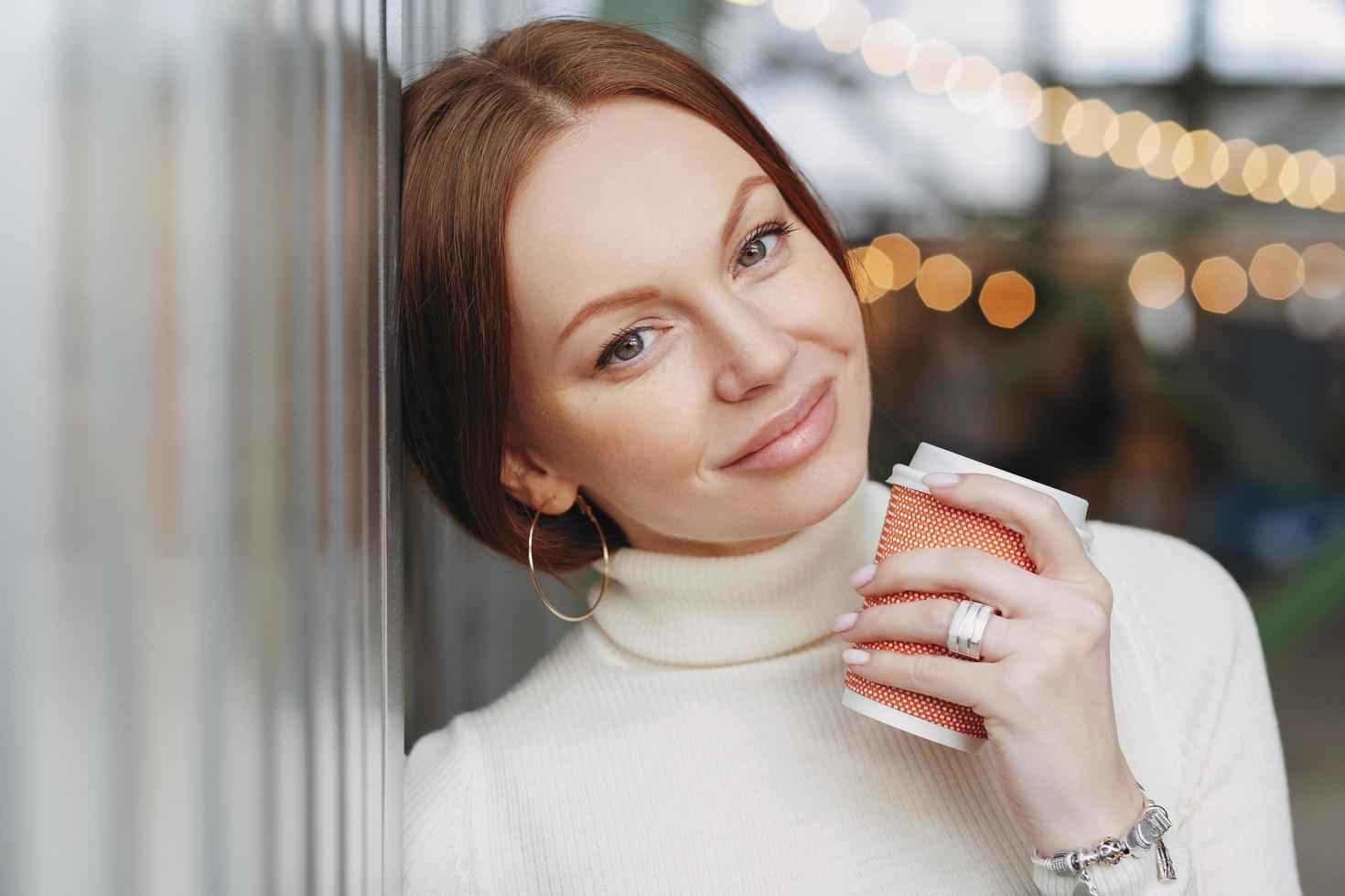 Dreamy pensive Caucasian woman with make up, dressed in casual outfit, leans on wall, holds takeaway coffee, remembers pleasant moments during outdoor stroll, has appealing look. Drink concept photo