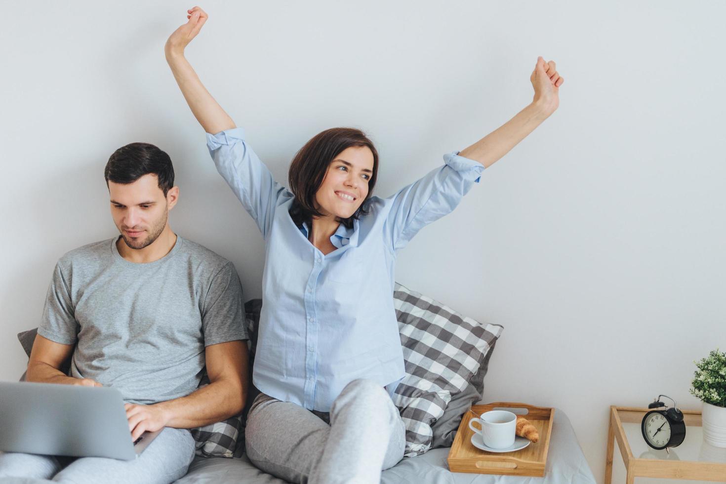 Smiling beautiful woman stretches in bed, enjoys good morning and being near her lover who works on modern laptop computer, happy to recieve coffee in bed, going to prepare breakfast. photo