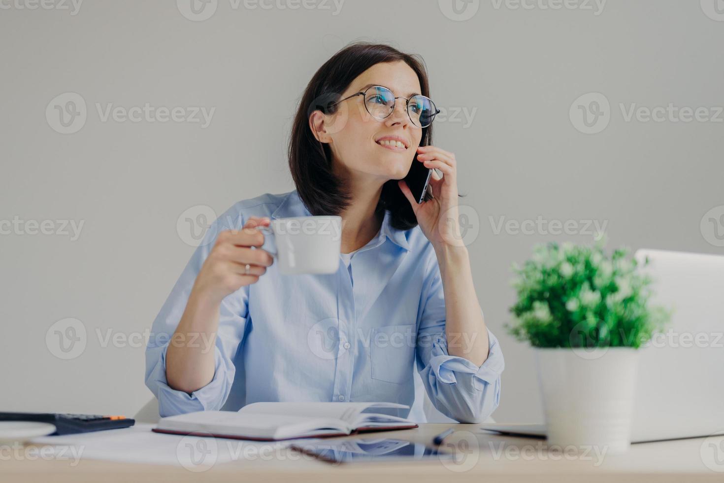 Gorgeous pleased businesswoman has cell phone conversation while has coffe break after working on notebook, shares her success with close person, isolated over grey background. Occupation concept photo