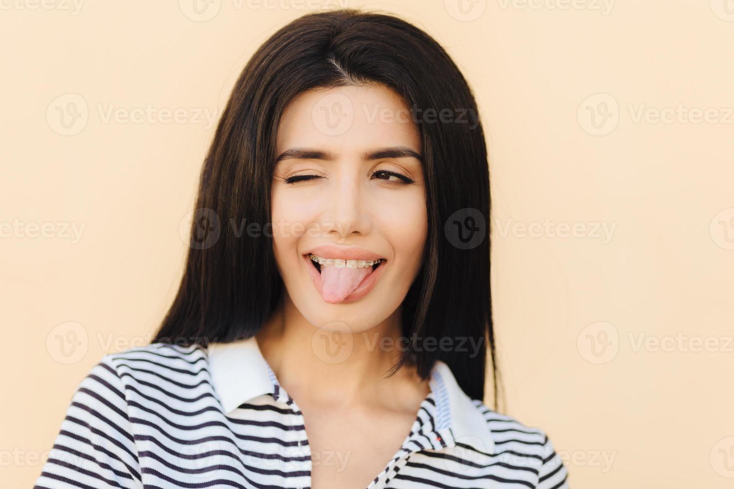 Funny joyful brunette female shows tongue, blinks with eye, makes grimace, wears braces, has long straight dark luxurious hair, dressed casually, isolated over light background. People and happiness photo