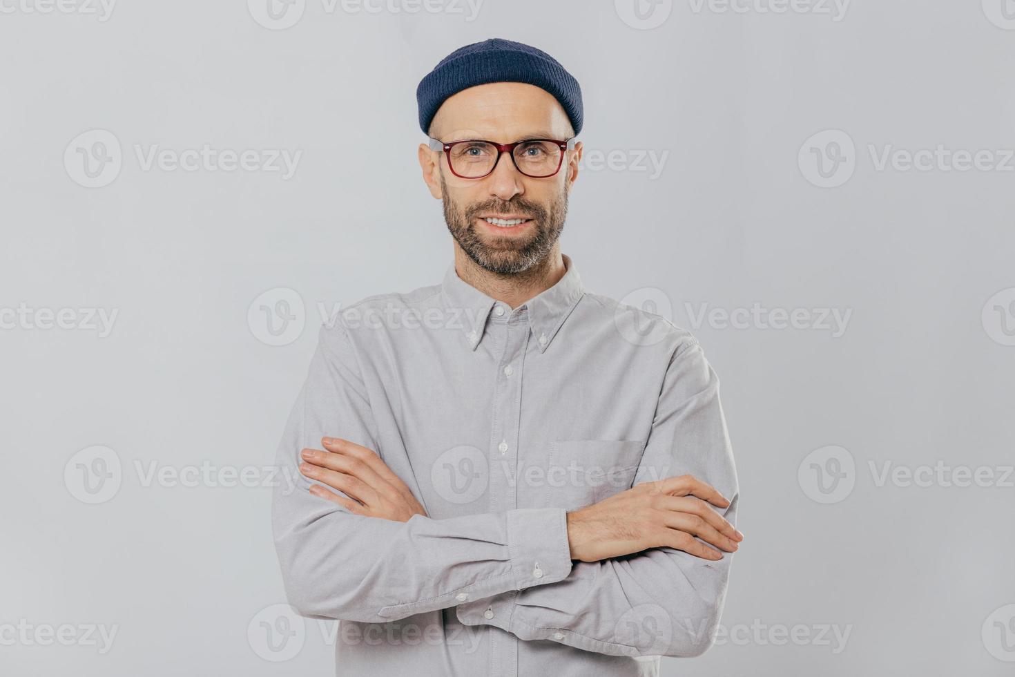 Horizontal shot of satisfied self confident male designer wears stylish headgear, dressed in white shirt, keeps arms folded, has dark stubble, isolated over white background. People and style concept photo